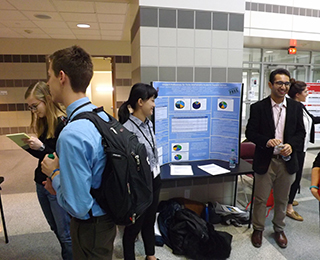 Photo of students in front of their poster with additional students in the background engaged in a discussion.